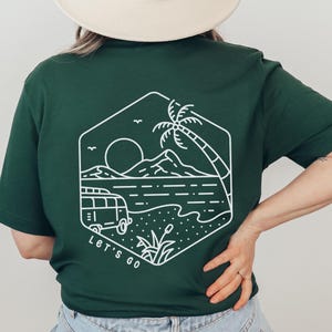 Op de afbeelding: Donkergroen t-shirt met een witte afbeelding van een strandscène, inclusief een bus, palmboom en zon. De tekst "Let's Go" staat eronder. De persoon draagt een crèmekleurige hoed en een blauwe jeans.
