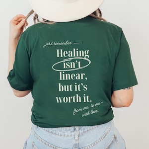 May include: A dark green t-shirt with the text "Healing isn't linear, but it's worth it." in white. The shirt also has the phrases "just remember" and "from me, to me - with love." A person is wearing the shirt and a white hat.
