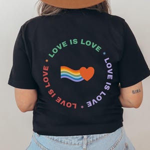 Könnte beinhalten: Schwarzes T-Shirt mit einem Regenbogen- und Herz-Motiv und dem Text "LOVE IS LOVE" in einem Kreis um das Motiv.