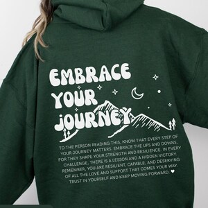 Puede incluir: Sudadera con capucha verde bosque con un estampado gráfico blanco que dice "Embrace Your Journey". El gráfico presenta montañas, estrellas y una luna creciente. El texto debajo del gráfico dice "A la persona que lee esto, sepa que cada paso de su viaje importa. Abrace los altibajos, porque moldean su fuerza y ​​resiliencia. En cada desafío, hay una lección y una victoria oculta. Recuerde, usted es resiliente, capaz y merece todo el amor y el apoyo que le llega. Confíe en sí mismo y siga adelante."