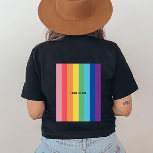 Könnte beinhalten: Schwarzes T-Shirt mit einem vertikalen Streifendesign in Regenbogenfarben auf dem Rücken. Das Design enthält den Text "LOVE IS LOVE" in Weiß. Die Person auf dem Bild trägt einen braunen Hut.