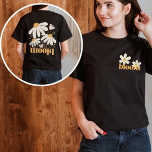 May include: Black t-shirt with a floral design and the word "bloom" in yellow. The front of the shirt features two white daisies with yellow centers and the word "bloom". The back of the shirt has a larger floral design with the same text.