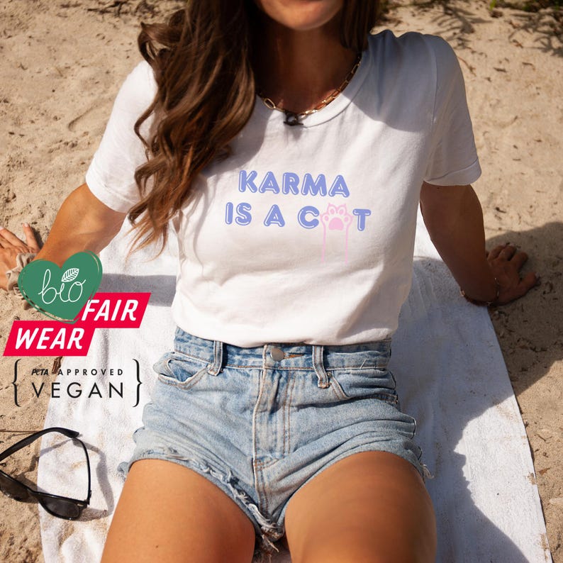 May include: A white t-shirt with the text "Karma is a cat" in blue and pink lettering. The text is surrounded by a pink paw print. The model is wearing the t-shirt with denim shorts and is sitting on a white towel on a sandy beach.
