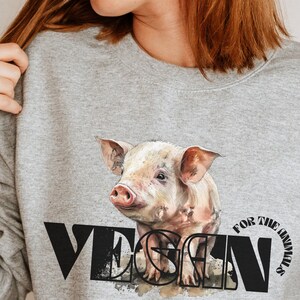 Op de afbeelding: Een grijs sweatshirt met een zwart-witte afbeelding van een varken en het woord "VEGAN" met de tekst "FOR THE ANIMALS" aan de rechterkant.