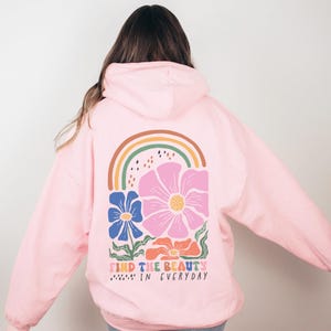 May include: A light pink hoodie with a floral and rainbow design on the back. The design includes a large pink flower, blue and orange flowers, and a rainbow. The text "FIND THE BEAUTY IN EVERYDAY" is printed below the flowers.
