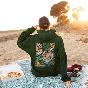 May include: Dark green hoodie with a floral design on the back. The design features large pink and yellow flowers with green leaves and the text "NO RAIN, NO FLOWERS". The hoodie is worn on a beach.