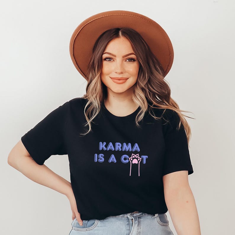 May include: A woman wearing a black t-shirt with the text "KARMA IS A CAT" in purple and blue lettering with a pink paw print. She is also wearing a brown fedora hat and blue jeans.