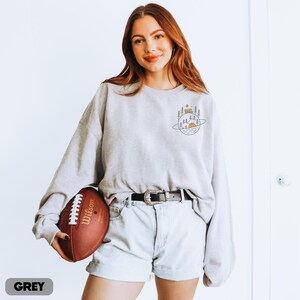 K&ouml;nnte beinhalten: Hellgraues Sweatshirt mit einem Camping-Motiv auf dem oberen R&uuml;cken. Das Sweatshirt wird mit hellblauen Jeansshorts und einem schwarzen G&uuml;rtel getragen. Ein brauner Football mit wei&szlig;en Schn&uuml;rsenkeln wird gehalten. Das Wort "GREY" steht unten.