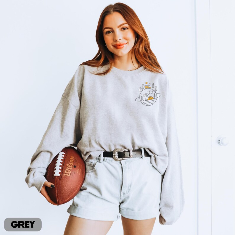 K&ouml;nnte beinhalten: Hellgraues Sweatshirt mit einem Camping-Motiv auf dem oberen R&uuml;cken. Das Sweatshirt wird mit hellblauen Jeansshorts und einem schwarzen G&uuml;rtel getragen. Ein brauner Football mit wei&szlig;en Schn&uuml;rsenkeln wird gehalten. Das Wort "GREY" steht unten.