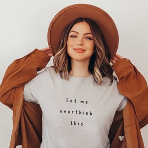Puede incluir: Una mujer con una camiseta gris con el texto "Let me overthink this" impreso en ella. Tambi&eacute;n lleva un sombrero marr&oacute;n y una chaqueta marr&oacute;n.