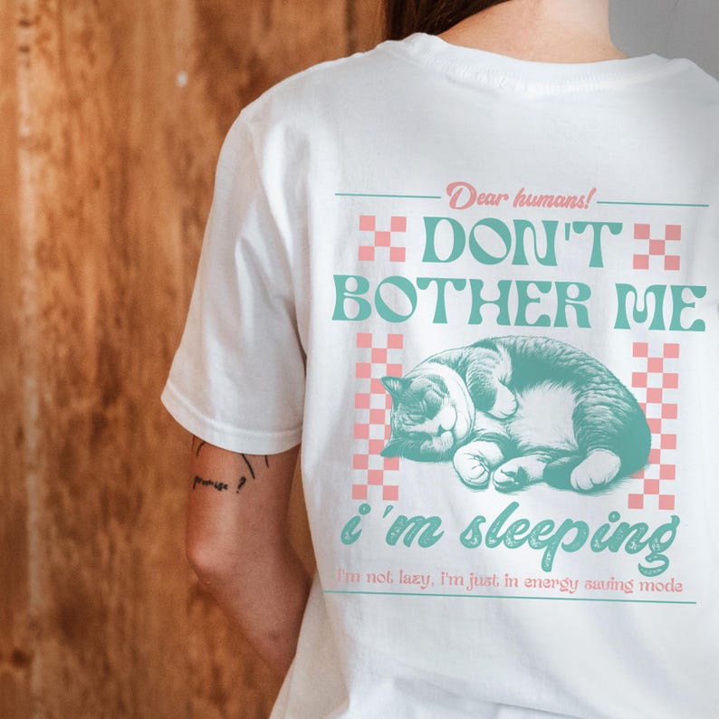 May include: White t-shirt with a graphic of a cat sleeping on a pink and white checkered background. The text "Dear humans! Don't bother me I'm sleeping I'm not lazy, I'm just in energy saving mode" is printed on the shirt.