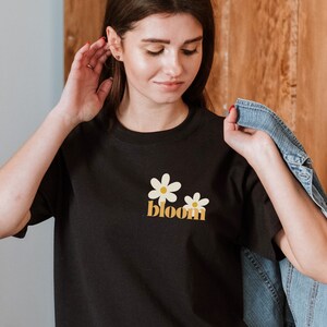May include: Black t-shirt featuring two white and yellow daisy flowers and the word "bloom" in a retro font. The shirt is a classic crew neck style.