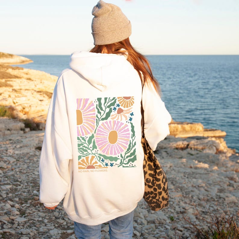 May include: White hoodie with a floral design on the back. The design features large pink and yellow flowers with green leaves and the text "NO RAIN, NO FLOWERS." The hoodie has a drawstring hood and long sleeves.