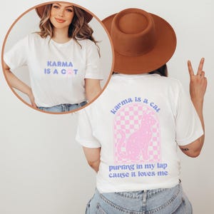 May include: White t-shirt with the text "KARMA IS A COT" on the front and "karma is a cat purring in my lap cause it loves me" on the back. The back design features a pink cat with white spots sitting on a pink and white checkered background.