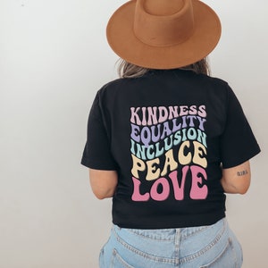 Könnte beinhalten: Schwarzes T-Shirt mit einem Retro-Grafikdesign mit den Worten "Kindness", "Equality", "Inclusion", "Peace" und "Love" in einem Regenbogen-Farbschema.