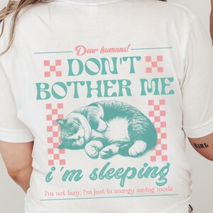May include: White t-shirt with a pink and blue graphic of a cat sleeping with the text "Dear humans! Don't bother me I'm sleeping I'm not lazy, I'm just in energy saving mode."