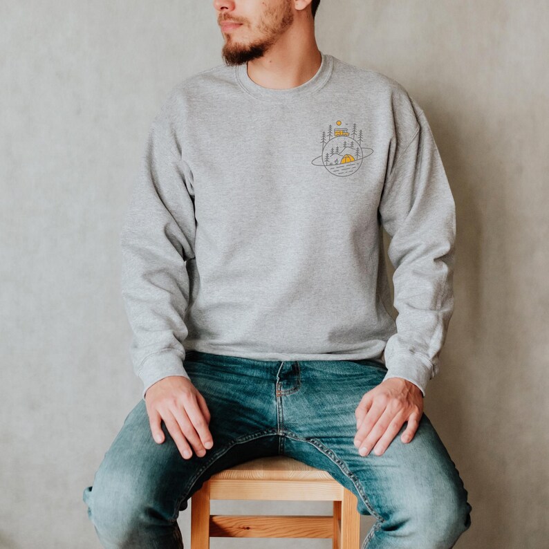 K&ouml;nnte beinhalten: Ein hellgraues Sweatshirt mit einer kleinen Camping-Grafik auf der Brust. Die Grafik zeigt ein Zelt, B&auml;ume und eine Sonne, alles innerhalb einer kreisf&ouml;rmigen Umrandung. Das Sweatshirt wird mit einer blauen Jeans kombiniert.