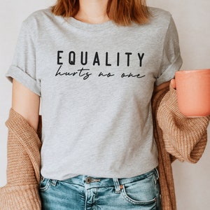 Könnte beinhalten: Hellgraues T-Shirt mit dem schwarzen Aufdruck "EQUALITY hurts no one". Das Shirt wird mit einer blauen Jeans und einer beigen Strickjacke getragen. Eine Person hält eine pfirsichfarbene Tasse.