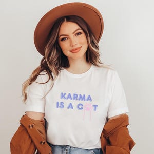 May include: A white t-shirt with the text "KARMA IS A CAT" in purple and a pink paw print. The model is wearing a brown hat and a brown jacket.
