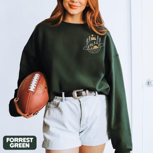K&ouml;nnte beinhalten: Forstgr&uuml;nes Sweatshirt mit einem Camping-Motiv auf der Brust. Das Design zeigt ein Zelt, B&auml;ume und eine Sonne, vor einem planeten&auml;hnlichen Hintergrund. Das Sweatshirt wird mit hellen Shorts und einem braunen Football kombiniert.