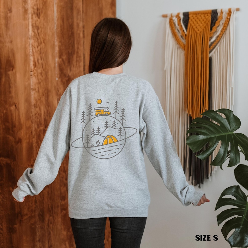 K&ouml;nnte beinhalten: Hellgraues Sweatshirt mit Camping-Motiv auf dem R&uuml;cken. Das Design zeigt einen gelben Camper, B&auml;ume, ein Zelt und ein Lagerfeuer in einer planeten&auml;hnlichen Umrandung. Das Sweatshirt ist Gr&ouml;&szlig;e S.