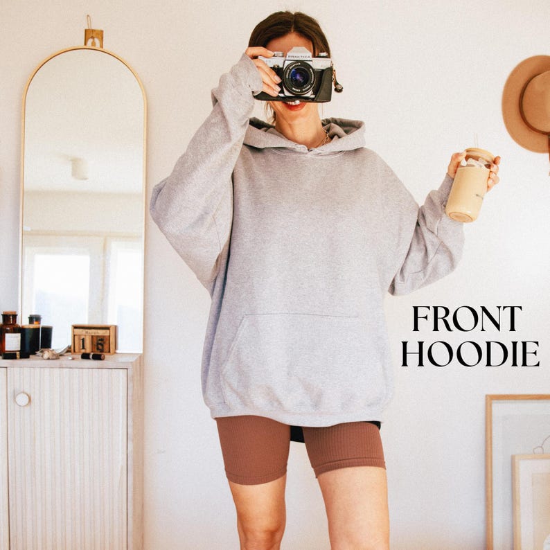 May include: A light grey hoodie with a front pocket, worn with brown biker shorts. The image also shows a vintage camera and a drink in a reusable cup. The text "FRONT HOODIE" is displayed in a bold font.