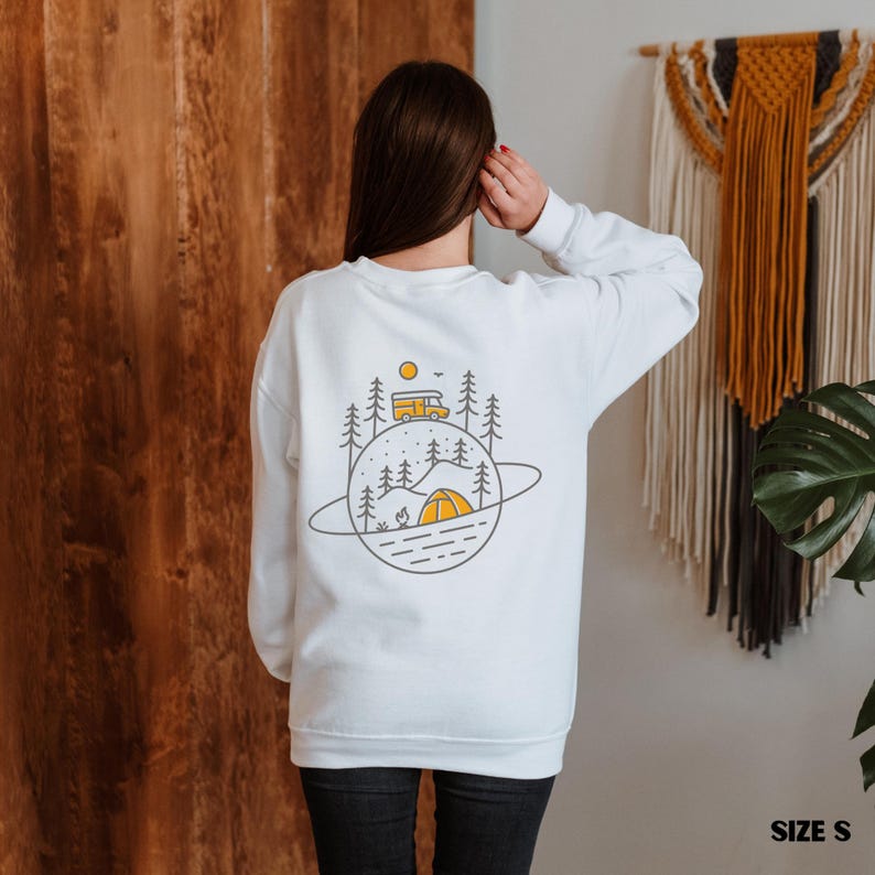 K&ouml;nnte beinhalten: Wei&szlig;es Sweatshirt mit Camping-Motiv auf dem R&uuml;cken. Das Design zeigt einen gelben Camper, ein Zelt, B&auml;ume und eine Sonne, alles innerhalb einer planetenartigen Umrandung. Das Sweatshirt ist Gr&ouml;&szlig;e S.