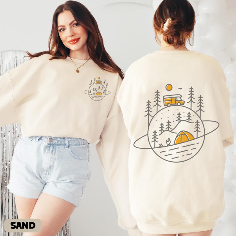 K&ouml;nnte beinhalten: Ein sandfarbenes Sweatshirt mit einem Camping-Motiv auf der Vorder- und R&uuml;ckseite. Das Design zeigt einen Camper, ein Zelt, B&auml;ume und ein Lagerfeuer innerhalb einer planetenartigen Umrandung. Das Wort "SAND" steht unten.