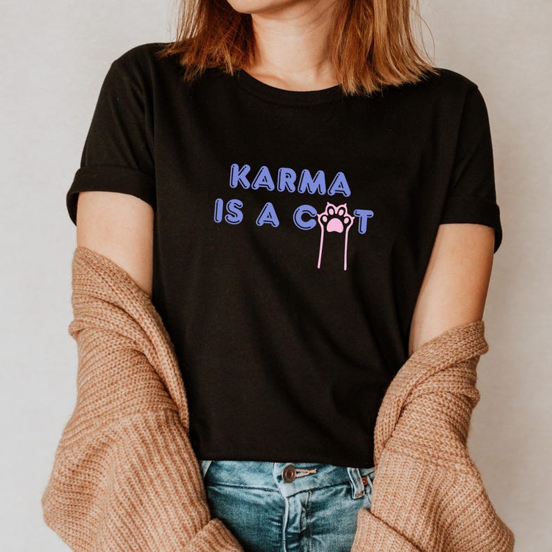 May include: A black t-shirt with the text "KARMA IS A CAT" in purple and pink lettering. The text includes a pink paw print.