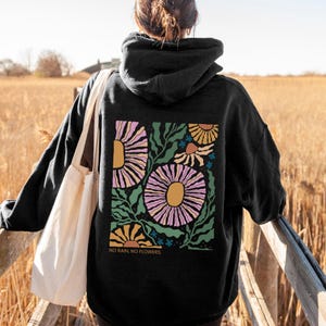May include: Black hoodie with a floral design on the back. The design features large, stylized flowers in shades of pink, yellow, and orange, with green leaves and the text "NO RAIN, NO FLOWERS." The person is wearing a white tote bag.