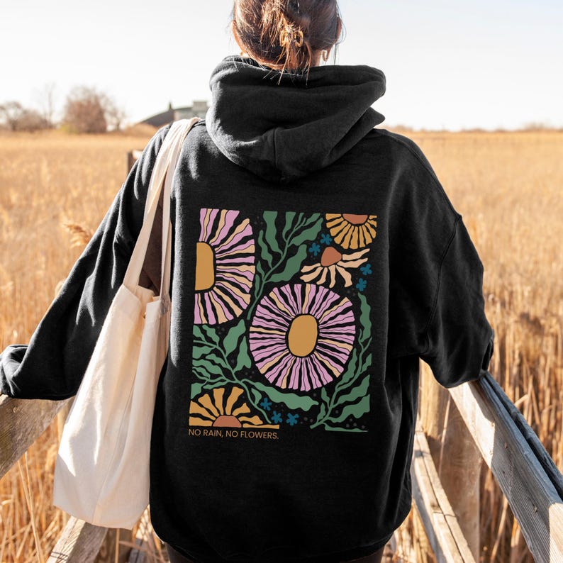 May include: Black hoodie with a floral design on the back. The design features large, stylized flowers in shades of pink, yellow, and orange, with green leaves and the text "NO RAIN, NO FLOWERS." The person is wearing a white tote bag.