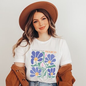Könnte beinhalten: Weißes T-Shirt mit einem Blumendesign und dem Text "Sei stolz auf deinen Fortschritt".