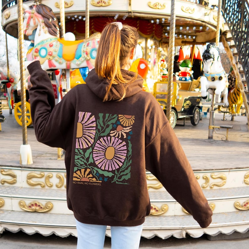 May include: A brown hoodie with a floral design on the back. The design features large pink and yellow flowers with green leaves and the text "NO RAIN, NO FLOWERS". The hoodie has a hood and long sleeves.