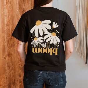 May include: Black t-shirt featuring a daisy floral design with the word "bloom" in a gold color. The daisy design has white petals and a yellow center. The shirt is a casual style.