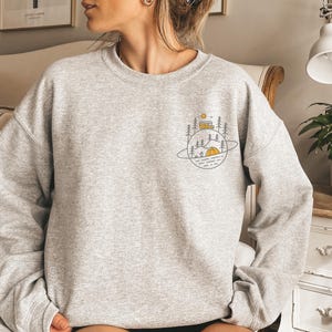 K&ouml;nnte beinhalten: Hellgraues Sweatshirt mit einem Camping-Motiv. Das Design zeigt ein gelbes Zelt, einen Wohnwagen, B&auml;ume und eine Sonne, alles innerhalb einer planeten&auml;hnlichen Umrandung. Das Sweatshirt ist aus weichem Material.