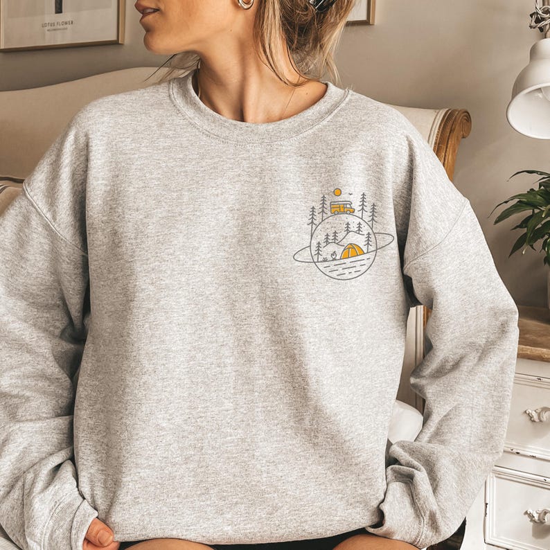 K&ouml;nnte beinhalten: Hellgraues Sweatshirt mit einem Camping-Motiv. Das Design zeigt ein gelbes Zelt, einen Wohnwagen, B&auml;ume und eine Sonne, alles innerhalb einer planeten&auml;hnlichen Umrandung. Das Sweatshirt ist aus weichem Material.