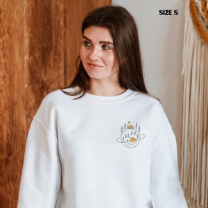 K&ouml;nnte beinhalten: Wei&szlig;es Sweatshirt mit einem Camping-Motiv. Das Design zeigt ein Zelt, B&auml;ume und einen Camper in einer planeten&auml;hnlichen Form. Der Text "SIZE S" ist oben rechts sichtbar.