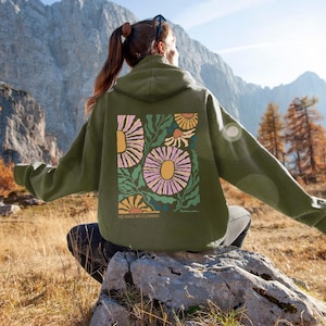 Può includere: Una felpa con cappuccio verde oliva con un design floreale sul retro. Il design presenta grandi fiori rosa e gialli con foglie verdi e il testo "NO RAIN, NO FLOWERS" in basso. La felpa con cappuccio è indossata all'aperto.