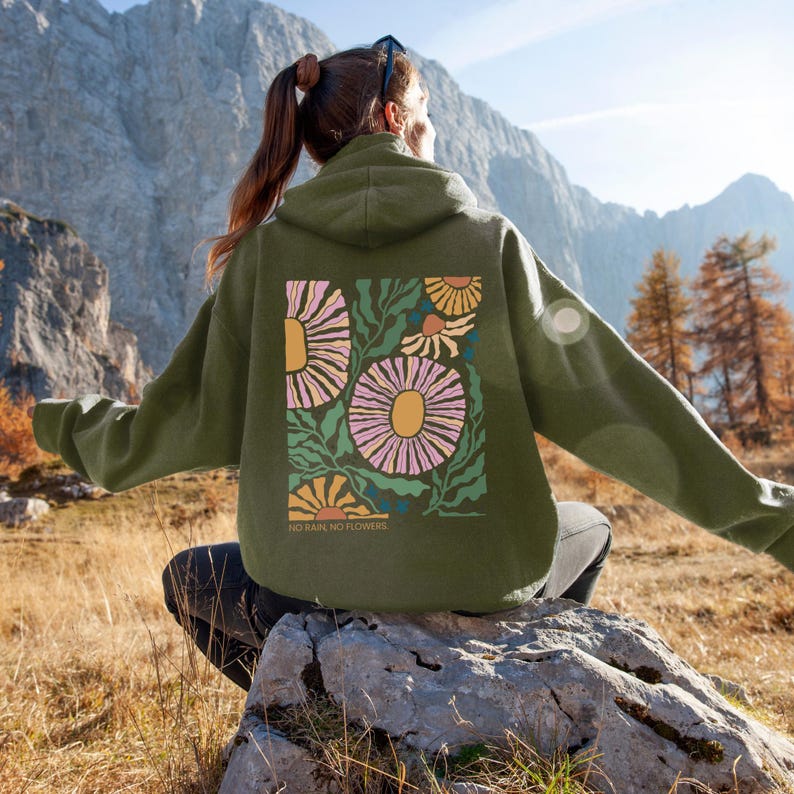 May include: An olive green hoodie with a floral design on the back. The design features large pink and yellow flowers with green leaves and the text "NO RAIN, NO FLOWERS". The hoodie is worn outdoors.