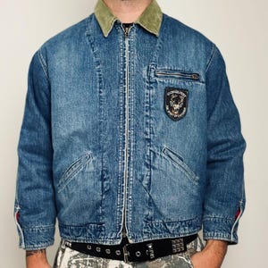 May include: A blue denim jacket with a light olive green corduroy collar and a front zipper closure. The jacket features a black patch on the left chest with a deer illustration and the text "CONQUEST THE SPIRIT OF CHEVIGNON" and a zippered pocket.
