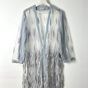 Pode incluir: Um cardigã longo aberto de cor azul claro transparente com um padrão de folhas prateadas em todo o tecido. A peça tem mangas compridas, um acabamento azul claro sólido ao longo da abertura frontal e um fecho decorativo prateado central.