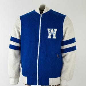 Pode incluir: Uma jaqueta estilo universitário azul e branca com textura felpuda. A jaqueta apresenta gola, punhos e bainha canelados em branco. Uma grande letra W branca com a palavra Wrangler impressa em azul está no peito esquerdo. Duas listras azuis acentuam cada manga.