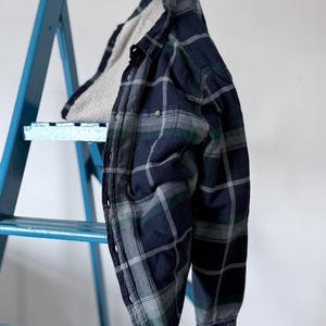 Könnte beinhalten: Ein langärmeliges Hemd mit Knopfleiste und einem Karomuster in Dunkelmarineblau, Waldgrün und Grau. Das Hemd ist mit einem dicken, strukturierten, cremeweißen Sherpa-Fleece-Material gefüttert, das am Kragen und an der vorderen Knopfleiste sichtbar ist.