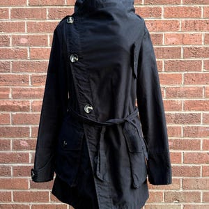 May include: A black jacket featuring an asymmetrical front closure with three visible buttons, a high collar, long sleeves with buttoned cuffs, and a tie belt at the waist. The jacket includes two large front pockets with flaps.