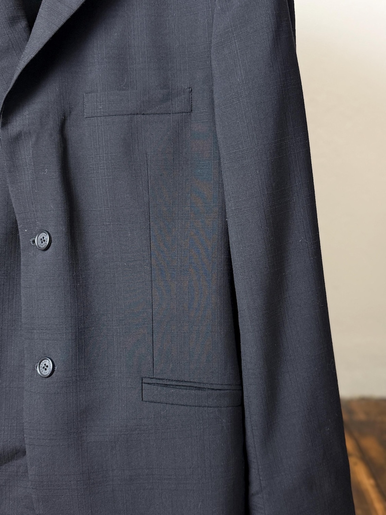 May include: A dark navy blue blazer with a subtle plaid pattern. The blazer features two buttons and a chest pocket. The fabric has a textured appearance. The blazer is a classic style.