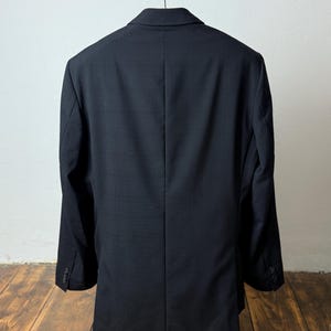 May include: A black blazer hanging on a metal hanger. The back of the blazer is visible, showing a tailored fit with a center vent. The blazer is made of a textured fabric and has buttoned cuffs. The blazer is displayed on a wooden stand.