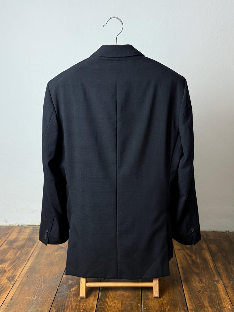 May include: A black blazer hanging on a metal hanger. The back of the blazer is visible, showing a tailored fit with a center vent. The blazer is made of a textured fabric and has buttoned cuffs. The blazer is displayed on a wooden stand.