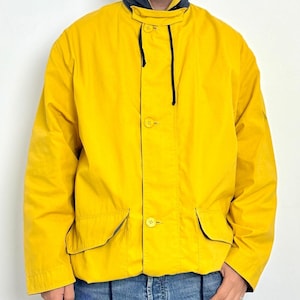 Può includere: Una giacca giallo brillante caratterizzata da un colletto alto con fodera interna blu scuro, chiusura frontale con bottoni e due ampie tasche con patta in vita. La giacca include un sottile cordino nero che pende dalla zona del colletto.