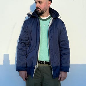 May include: A navy blue hooded jacket with a light-colored lining, worn over a light green crew-neck shirt and olive green cargo pants. The outfit includes a black beanie, a black belt with silver hardware, and a silver cross pendant necklace.