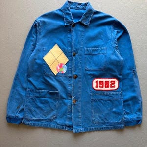 May include: A blue long-sleeve work jacket featuring a pointed collar, a button-down front, and four front pockets. The jacket includes a yellow diamond-shaped patch, a circular multi-colored pin, and a rectangular patch with the red text 1982.